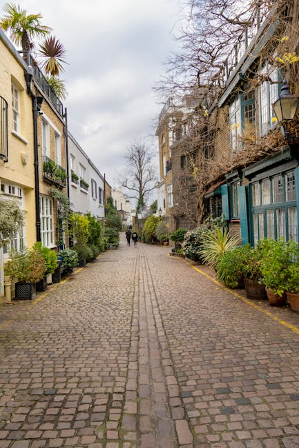 A quiet cobblestone street in a residential area with narrow pavement, flanked by colorful houses and garden spaces. On the left side, there are small, neatly maintained garden beds with lush green shrubs, potted plants, and flowering bushes, adding vibrant touches to the building facades. The right side features a mix of buildings with large windows and external plant displays, some with trellises or climbing plants. The street is lined with paving stones showing subtle textures, with a central groove for drainage. In the background, tall, leafless trees suggest early spring or late autumn, with overcast skies creating soft, diffused lighting. Gardeners South Kensington are pictured in the distance, walking along the street, amid a calm outdoor setting that emphasizes garden maintenance and landscaping work typical of a charming urban garden scene.