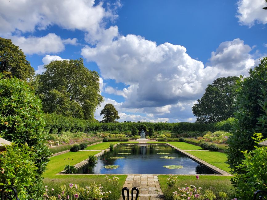 A panoramic view of a well-maintained formal garden featuring a rectangular pond at the center with a small fountain in the middle, surrounded by neatly trimmed lawn and flowerbeds with blooming plants. The garden is bordered by lush, green hedges and mature trees under a bright blue sky filled with fluffy white clouds. The grassy pathways are clean and appear recently mowed, and the scene suggests careful garden maintenance and landscaping work, possibly performed by professionals such as Gardeners South Kensington, to create a peaceful outdoor space suitable for lawn care, hedge trimming, and outdoor relaxation.
