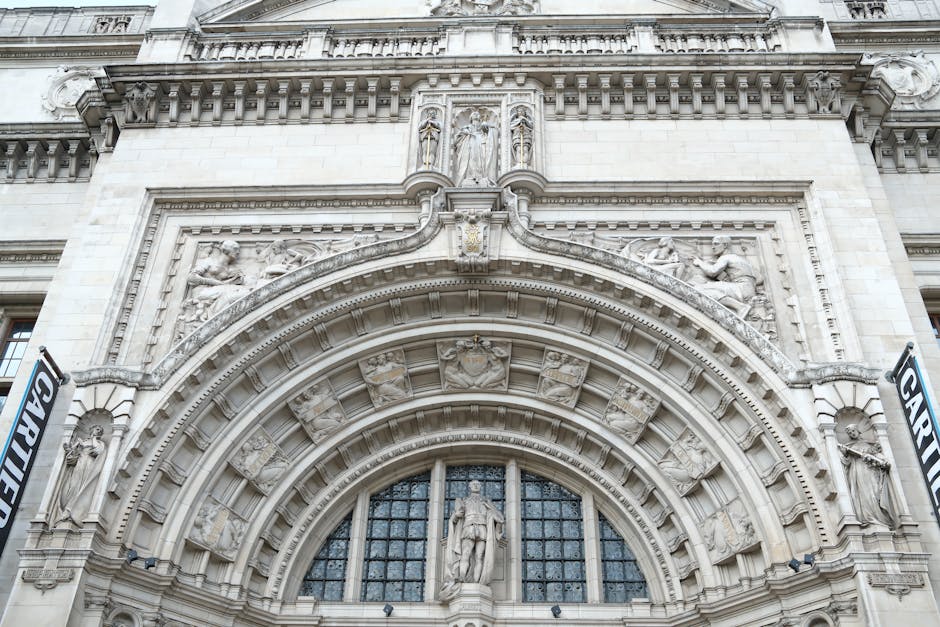 A detailed view of the ornate exterior of a historic building, featuring intricate stone carvings and sculptures around a large arched window. The façade displays classical architectural elements such as decorative moldings, statues, and reliefs, with a prominent central figure standing in the middle of the arch. The stone surface shows signs of weathering, highlighting its age and craftsmanship. Although the image focuses on the building's façade, the context suggests an outdoor setting with natural light illuminating the detailed stonework. This architectural style complements garden maintenance themes by emphasizing the craftsmanship involved in historic landscaping and exterior decoration, aligning with services offered by Gardeners South Kensington for high-quality outdoor and landscape care near Victoria and Albert Museum South Kensington.