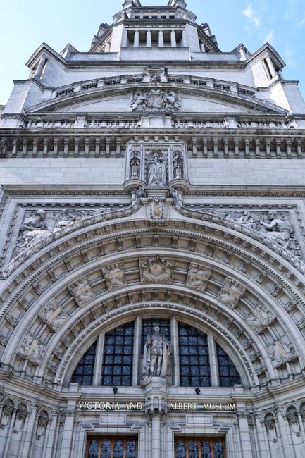 The image shows an upward view of the ornate facade of the Victoria and Albert Museum in South Kensington, showcasing detailed stone carvings and sculptures above the arched entrance. The architecture features intricate decorative elements, such as figures and floral motifs, with a large arched window framed by sculpted molding and reliefwork. The weather appears clear with blue sky visible at the top. This scene does not include any garden or landscaping elements, focusing solely on the historic building's detailed exterior architecture.