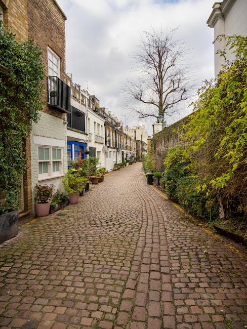 A narrow cobbled pathway in a residential garden setting, flanked by white terraced houses on the left with small balconies and potted plants, and a high stone wall covered with green foliage on the right. In the background, a leafless tree stands tall against a cloudy sky, providing a natural focal point amid the urban environment. The path shows signs of regular maintenance with clean, even cobblestones, and the surrounding plants appear well cared for. Gardeners South Kensington specializes in garden maintenance and landscaping work, ensuring tidy borders, lush greenery, and a pristine outdoor appearance. The scene conveys a quiet, well-kept outdoor space, ideal for street-level garden services and hedge trimming, blending urban charm with professional outdoor care.