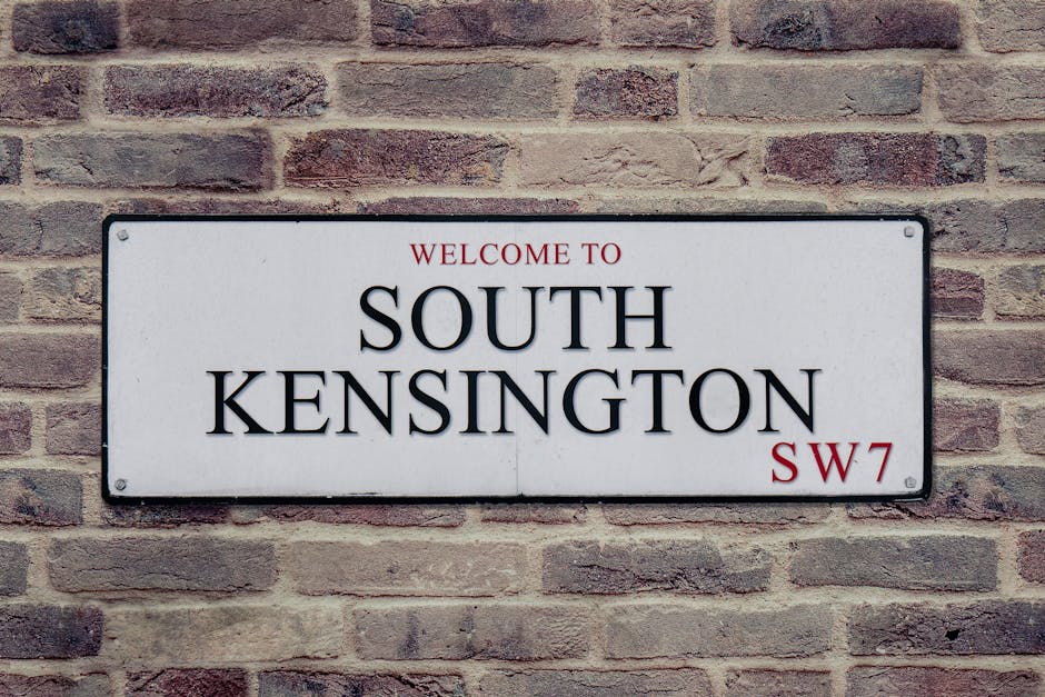 A rectangular white sign mounted on a brick wall displaying a welcoming message for South Kensington with the text 'WELCOME TO SOUTH KENSINGTON SW7' in black and red lettering. The brick wall has a textured surface with reddish-brown and grayish bricks, some with visible mortar. The sign appears clean and well-maintained, suggesting professional garden maintenance or landscaping work by Gardeners South Kensington. The background highlights no outdoor elements but emphasizes the signage's placement on an exterior wall, hinting at the entrance to a garden or residential area in South Kensington.